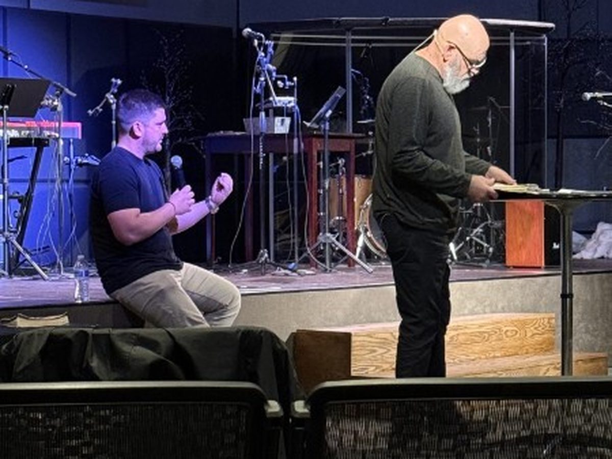 A young man kneeling in prayer while a leader reads scripture